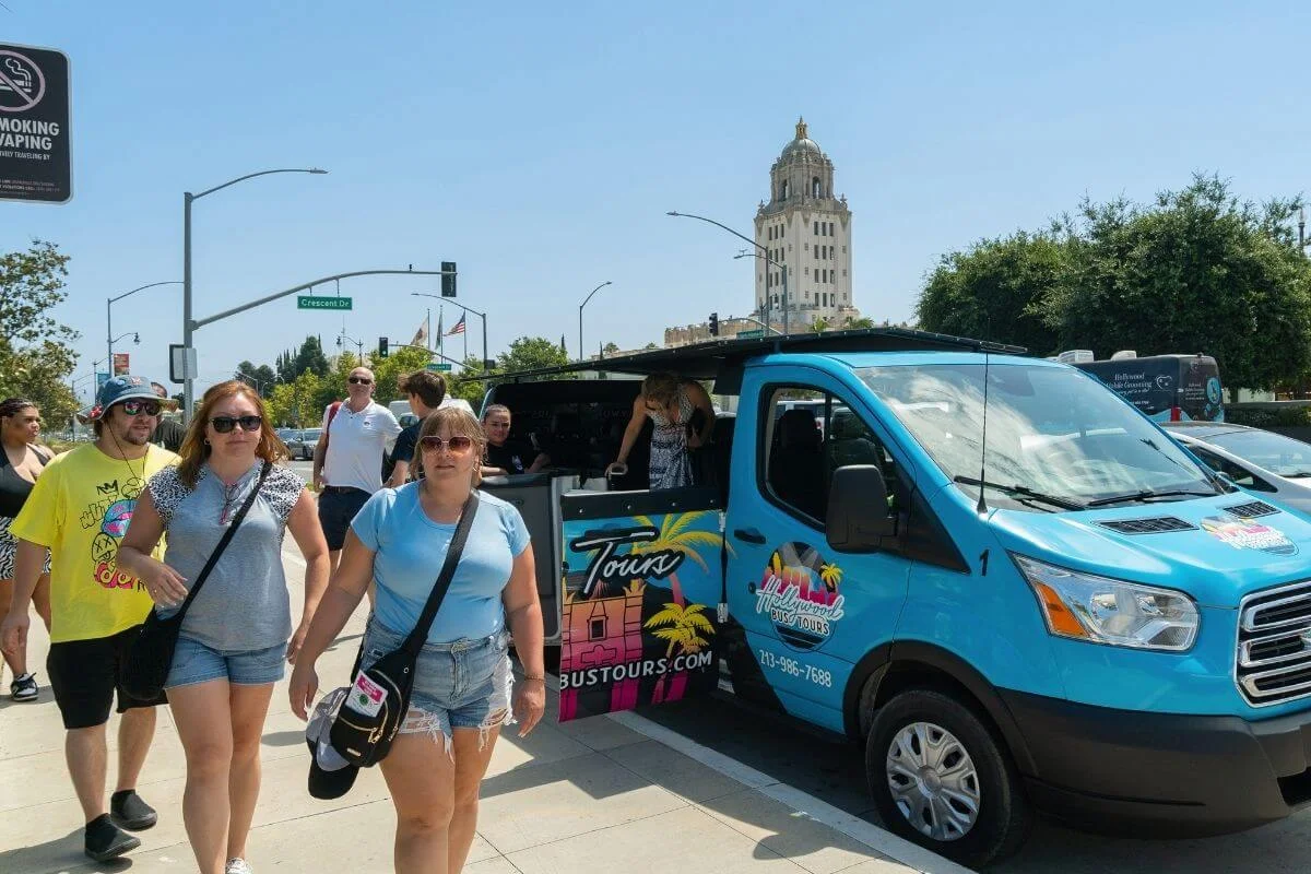 Tourists walk along a sunny sidewalk beside a bright blue Hollywood Bus Tours van with its side door open, while more people gather near the vehicle and a tall historic building rises in the background.