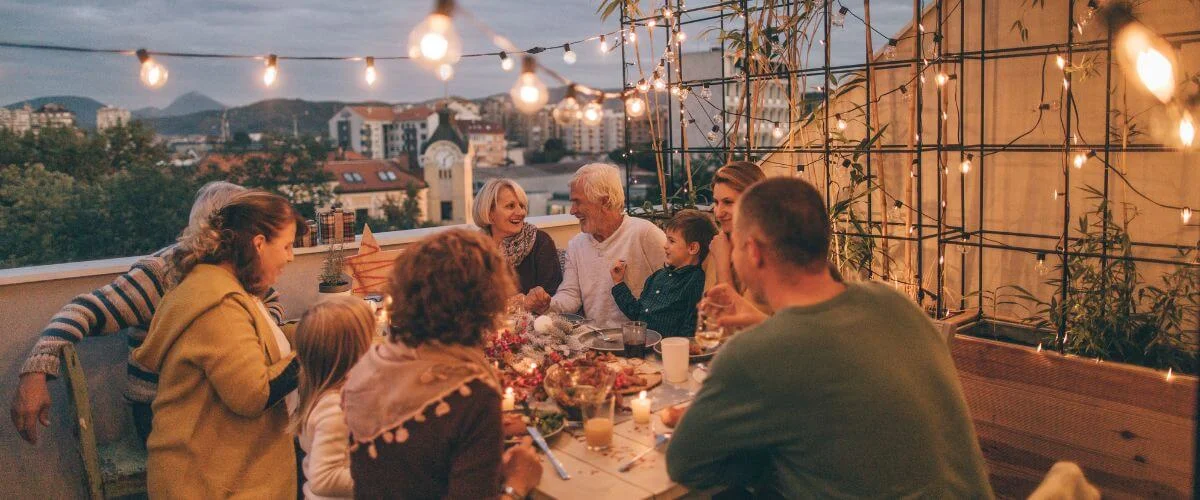 Multi-generational family shares a cozy evening meal on a rooftop terrace at dusk, with warm string lights and a city skyline in the background.
