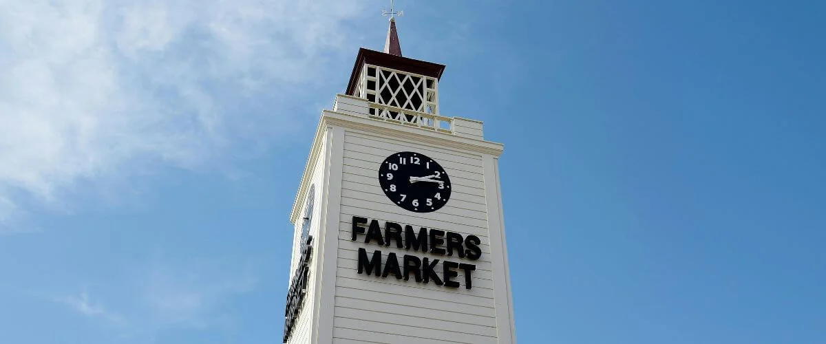Tall white clock tower with black “Farmers Market” lettering and a round clock near the top, standing against a bright, cloud-dotted blue sky.