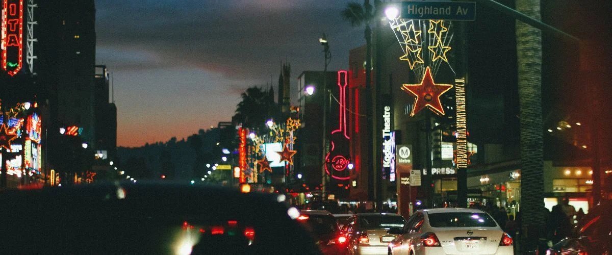 Evening view of cars in heavy traffic along Hollywood Boulevard near Highland Avenue, with neon signs, star-shaped lights, and palm trees glowing against a darkening sky.