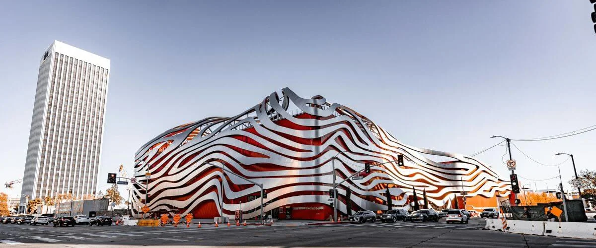 Wide street view of a modern museum with flowing red and silver metal ribbons covering its exterior, cars stopped at the intersection in front, and a tall office building in the background under a clear sky.