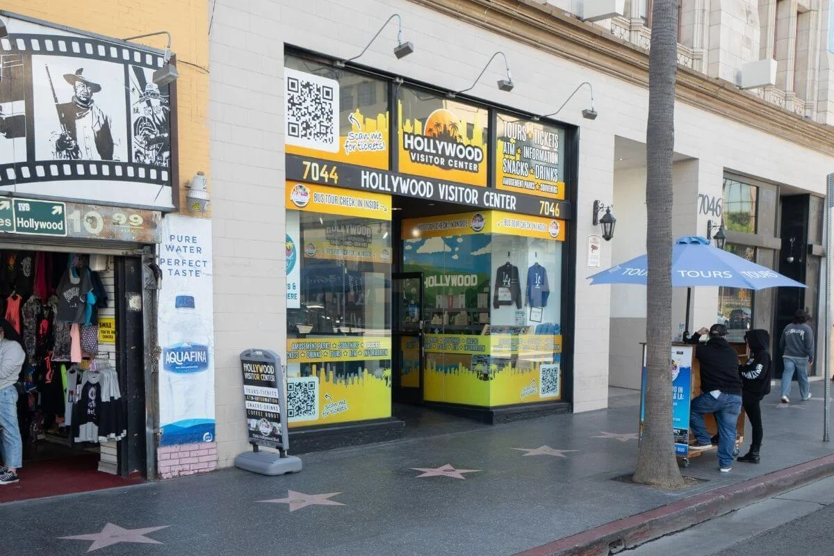 Street view of the Hollywood Visitor Center storefront with bright yellow signage, a nearby souvenir shop, tour kiosk with blue umbrella, and Hollywood Walk of Fame stars on the sidewalk in front.
