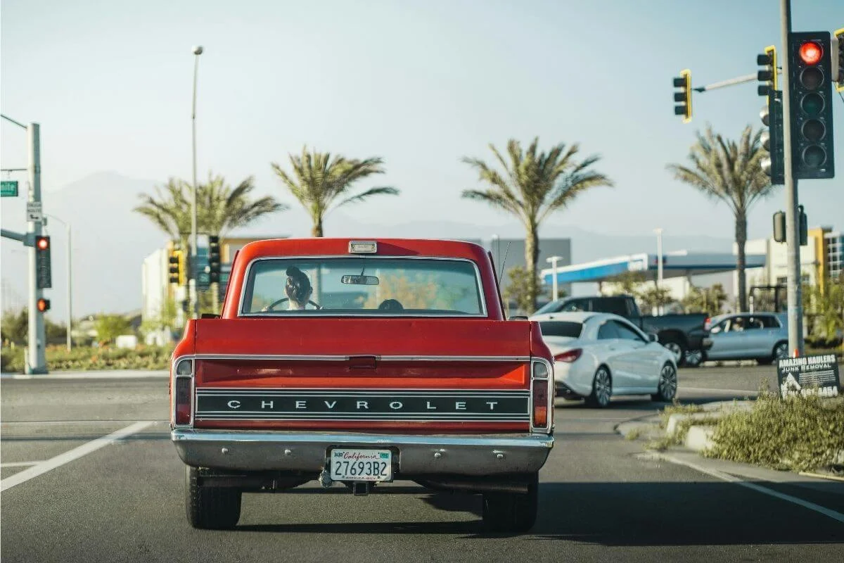 Rear view of a bright red vintage Chevrolet pickup truck stopped at a traffic light on a sunny day, with palm trees, other cars, and distant mountains in the background.
