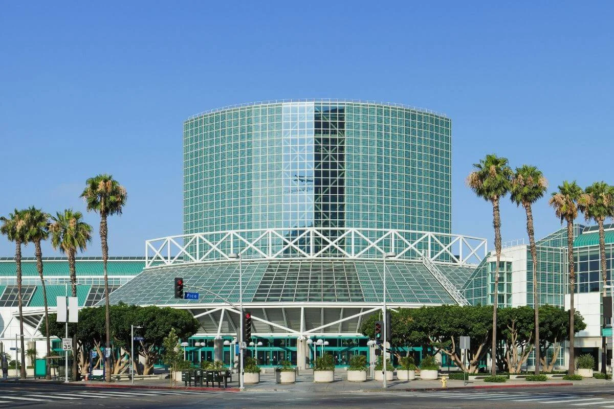 Exterior view of the Los Angeles Convention Center with its large glass cylinder tower, palm trees, and entrance structure under a clear blue sky.
