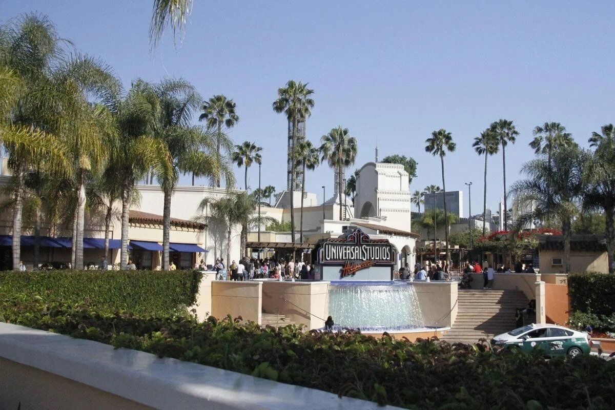 Visitors gather near the Universal Studios Hollywood entrance sign and fountain, surrounded by palm trees and buildings on a sunny day.