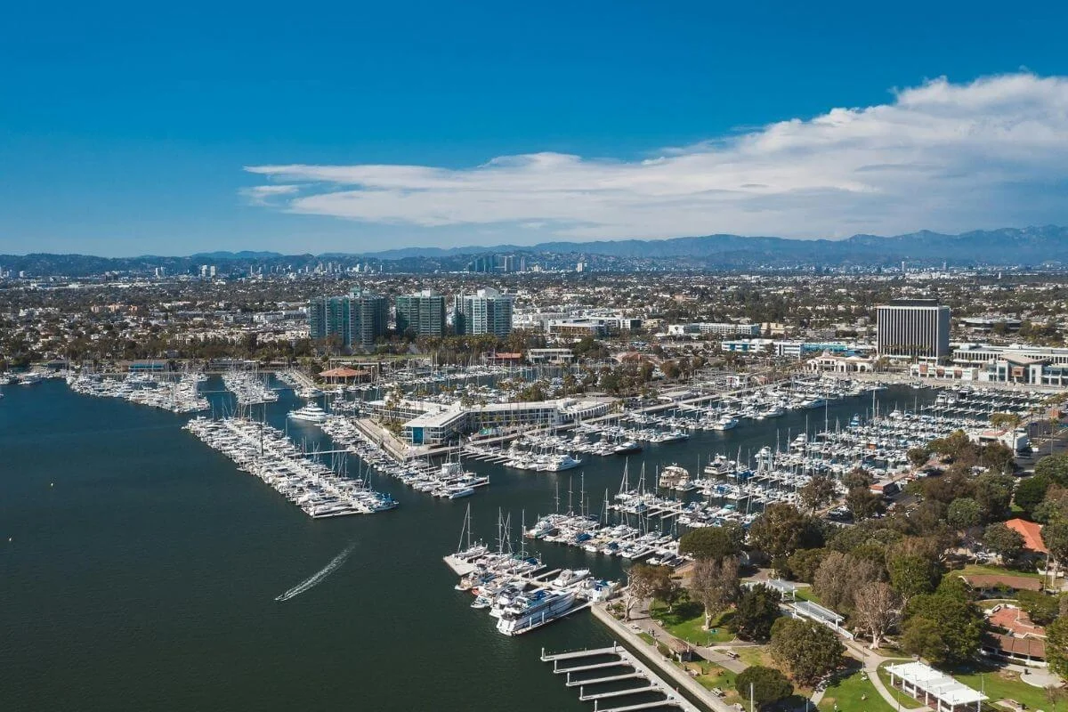 Drone view of a large marina packed with sailboats and yachts along the waterfront, with city buildings and distant mountains under a bright blue sky.