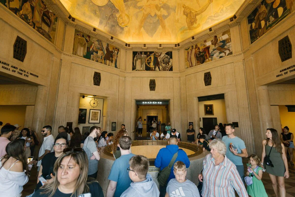 A large group of visitors fills a circular museum hall with a central exhibit, surrounded by tall stone walls and colorful murals near the ceiling.