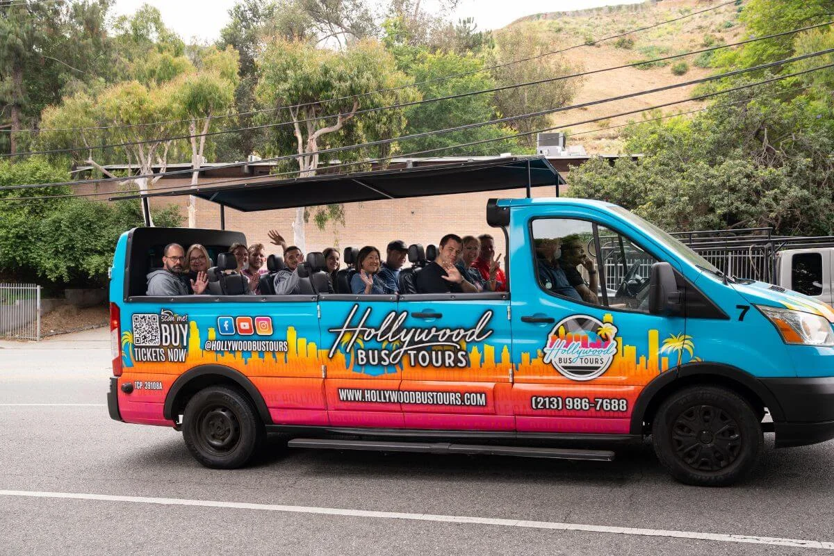A bright blue and orange open-air Hollywood Bus Tours vehicle filled with smiling passengers waves at the camera while driving along a tree-lined street.