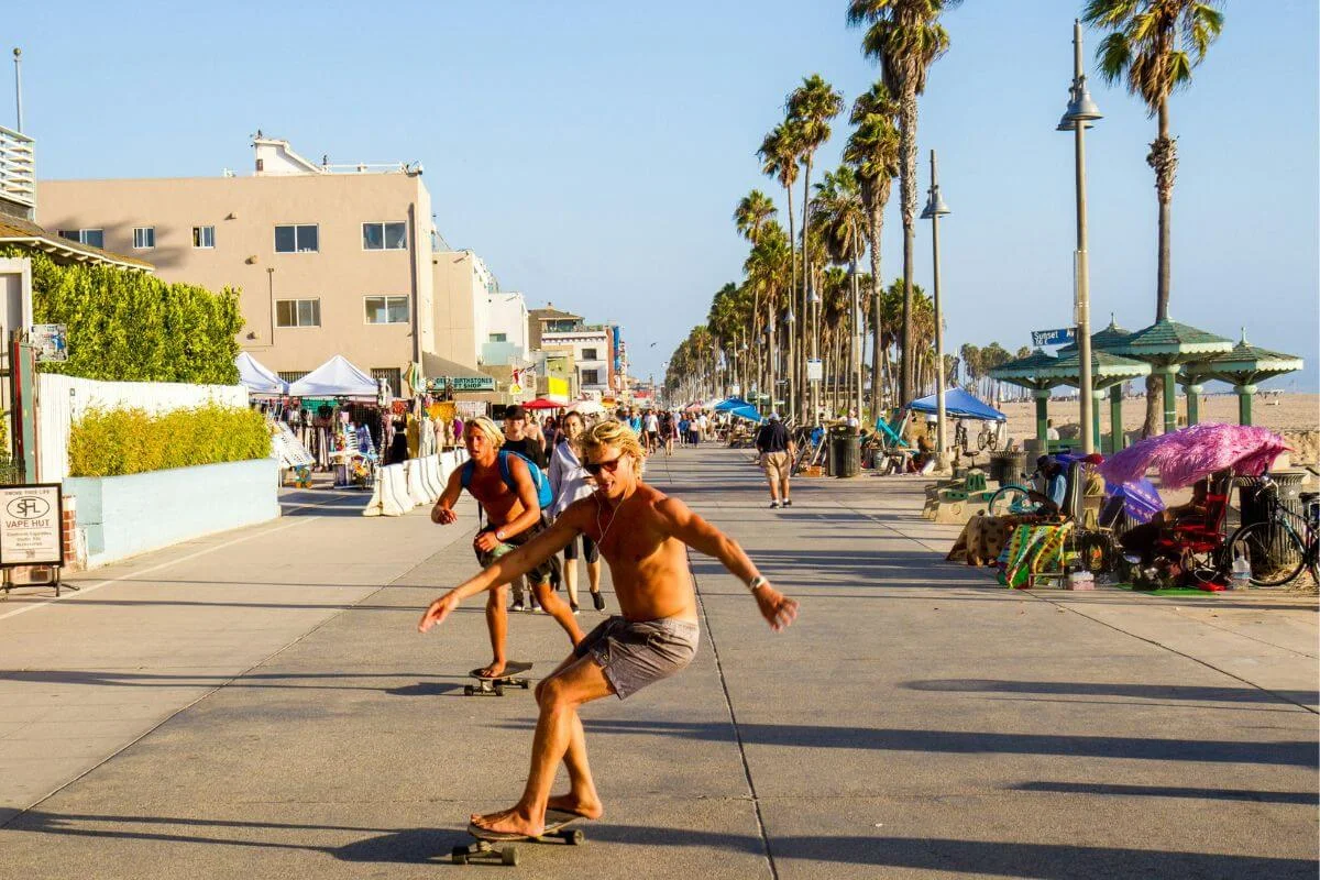Two shirtless skateboarders ride along the busy Venice Beach boardwalk on a sunny day, passing palm trees, vendors, and pedestrians with the sandy beach visible to the right.