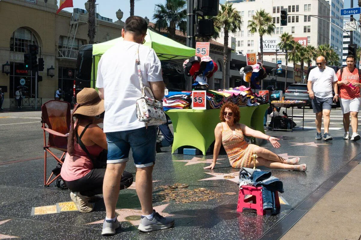 A woman poses sitting on the Hollywood Walk of Fame stars while another person photographs her, with street vendors selling $5 hats and shirts under a green tent and pedestrians walking past palm-lined buildings in the background.
