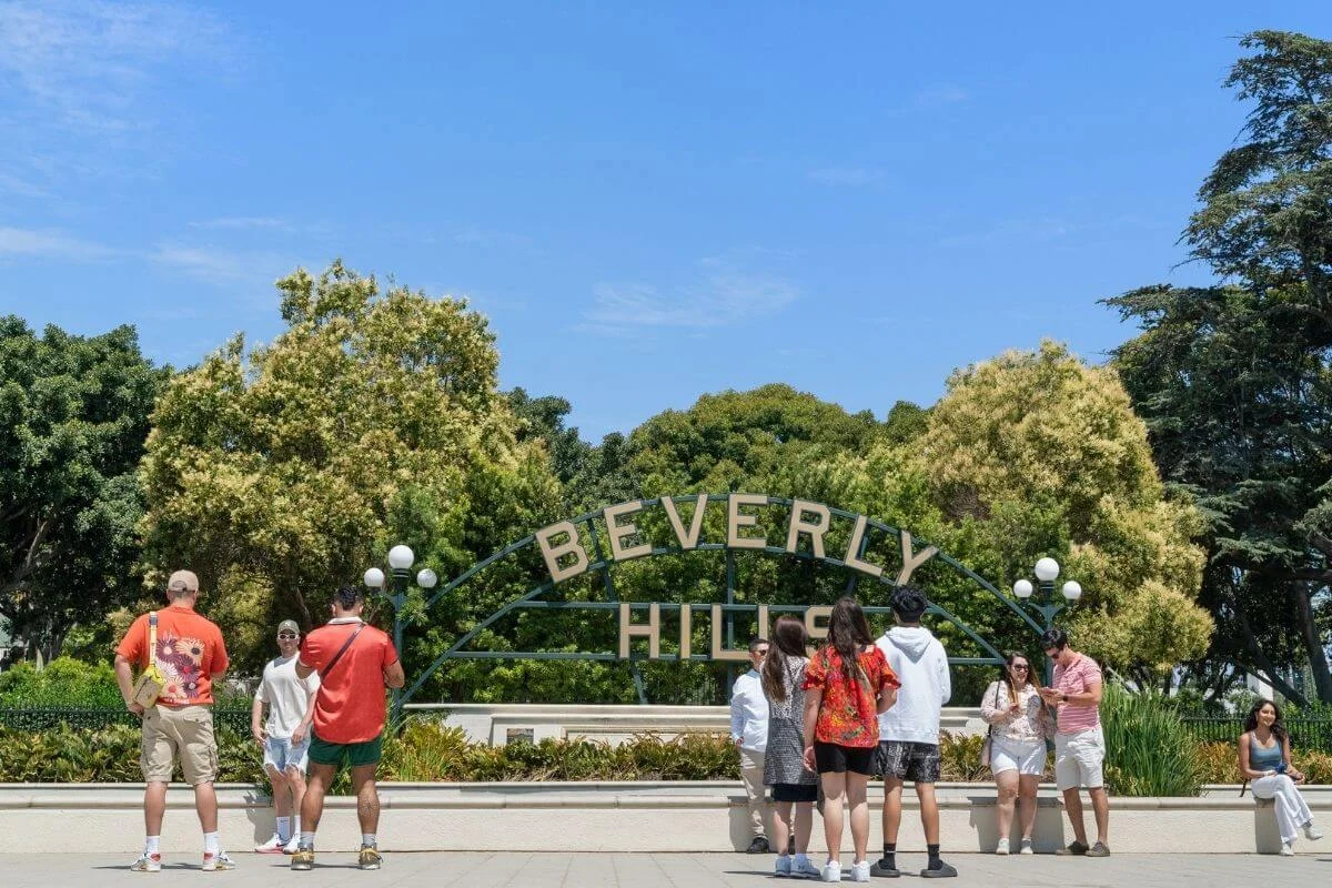 A group of people standing and taking photos in front of the iconic Beverly Hills sign, surrounded by lush green trees under a clear blue sky.