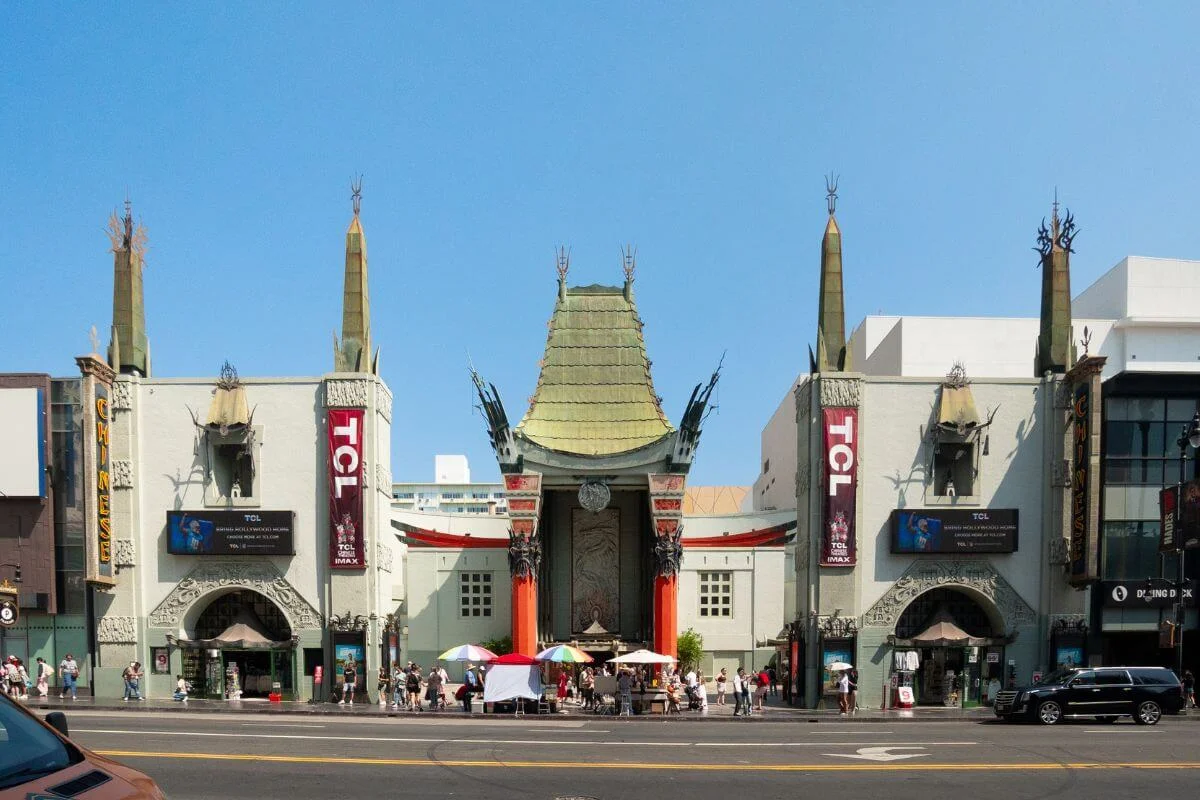 Street view of the ornate TCL Chinese Theatre in Hollywood, with its green pagoda-style roof, tall spires, and red columns, as tourists and street vendors gather on the sidewalk in front.