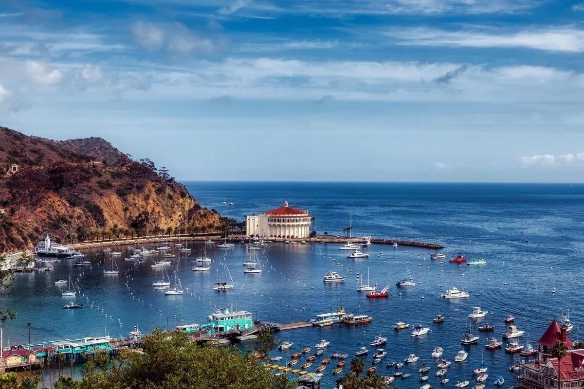 An aerial view of Avalon Harbor on Catalina Island, showing boats anchored in clear blue water, a round waterfront building near the shoreline, surrounding hills, and the open ocean beyond under a partly cloudy sky.