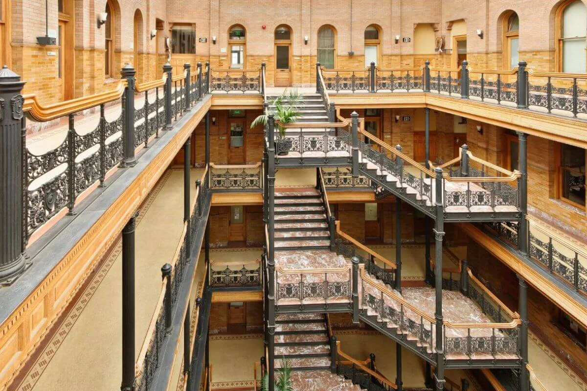Interior view of a multi-story atrium featuring decorative black iron railings, wide marble staircases, and warm brick walls with wooden doors lining each level.