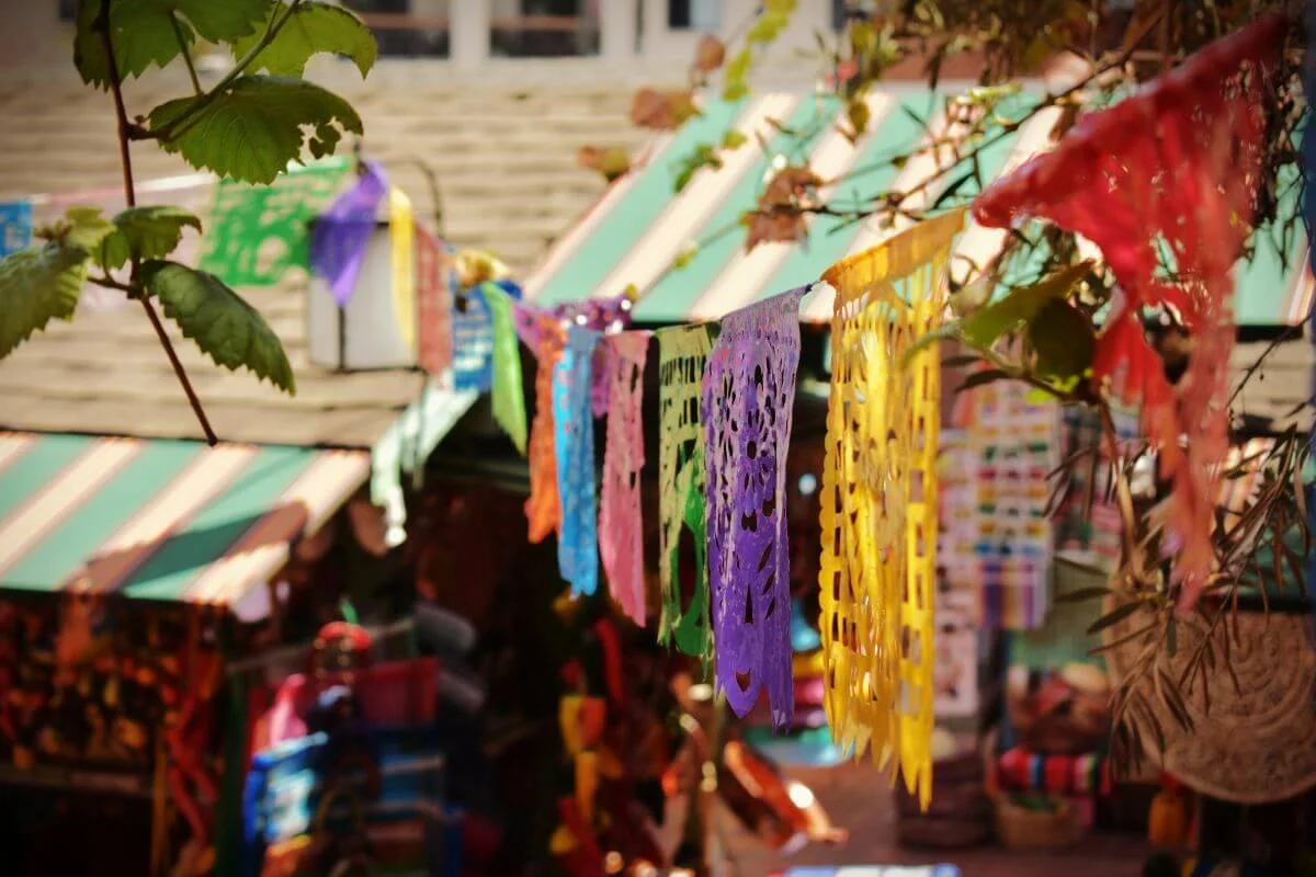 Brightly colored papel picado banners hang across an outdoor market with striped awnings, leafy branches, and stalls filled with vibrant crafts and textiles in the background.