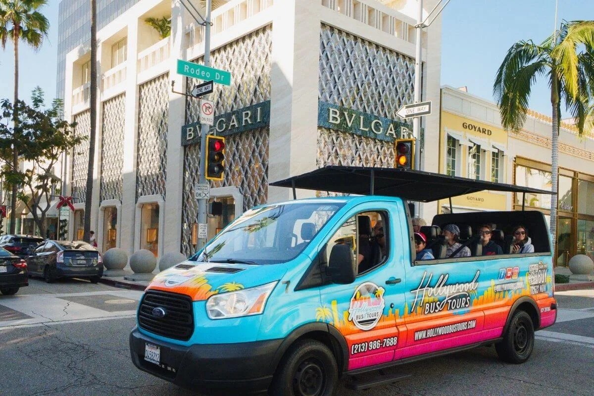 A colorful Hollywood sightseeing bus driving past luxury storefronts on Rodeo Drive in Beverly Hills, with passengers seated inside and designer brand buildings in the background.