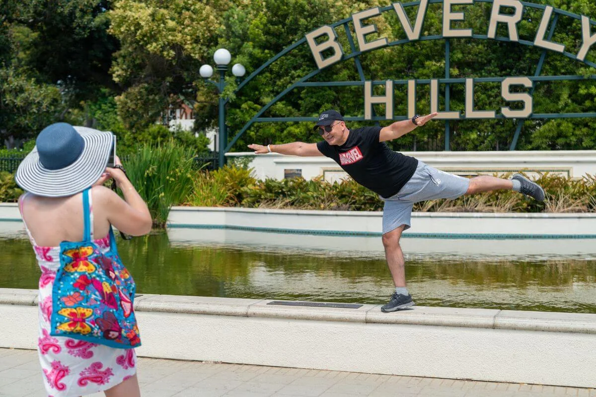 A man playfully balances on one leg along the edge of a fountain in front of the Beverly Hills sign while a woman in a sunhat and colorful beach cover-up takes his photo.