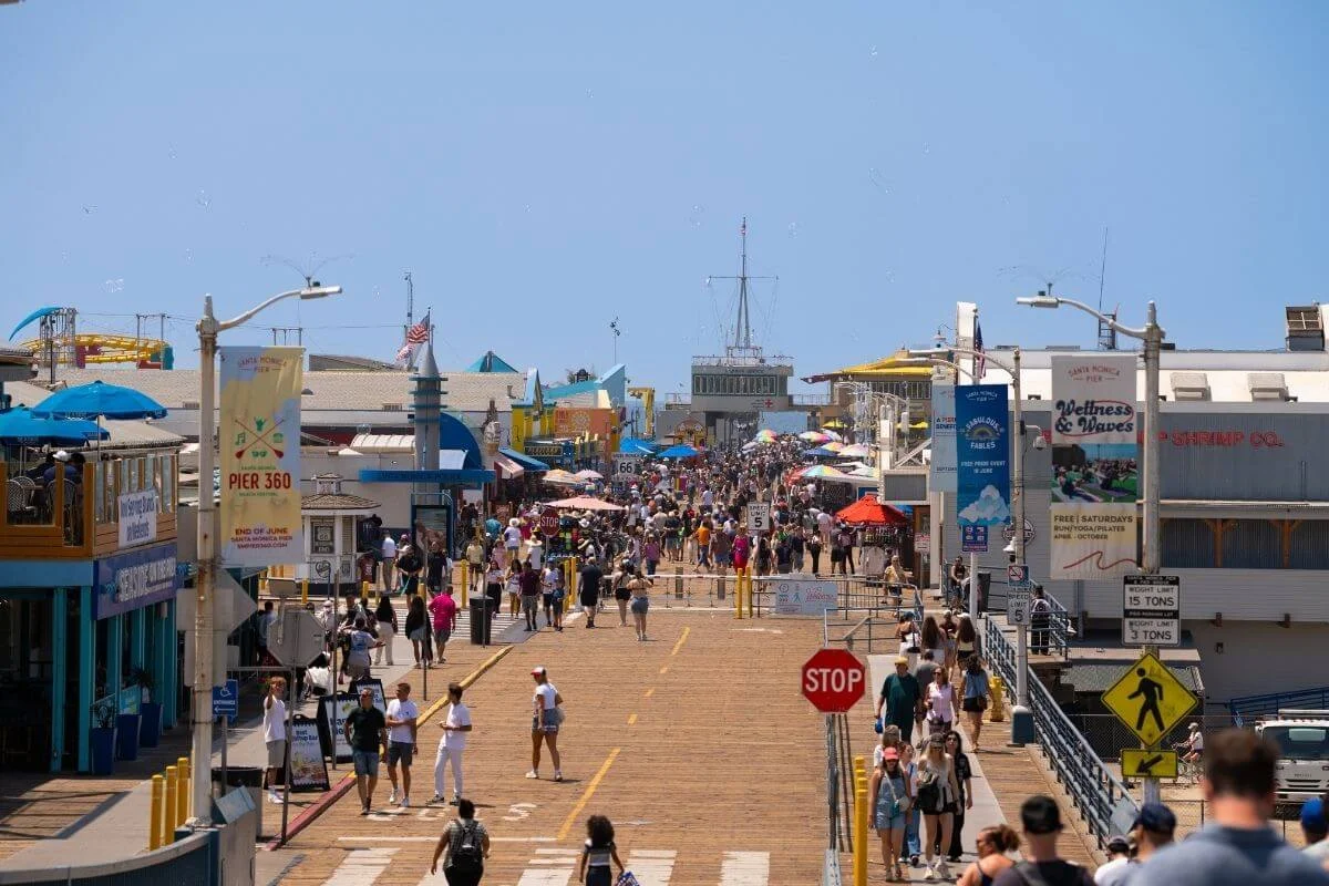 A busy view of the Santa Monica Pier with crowds of people walking along the wooden boardwalk, surrounded by shops, attractions, and signs under a clear blue sky.