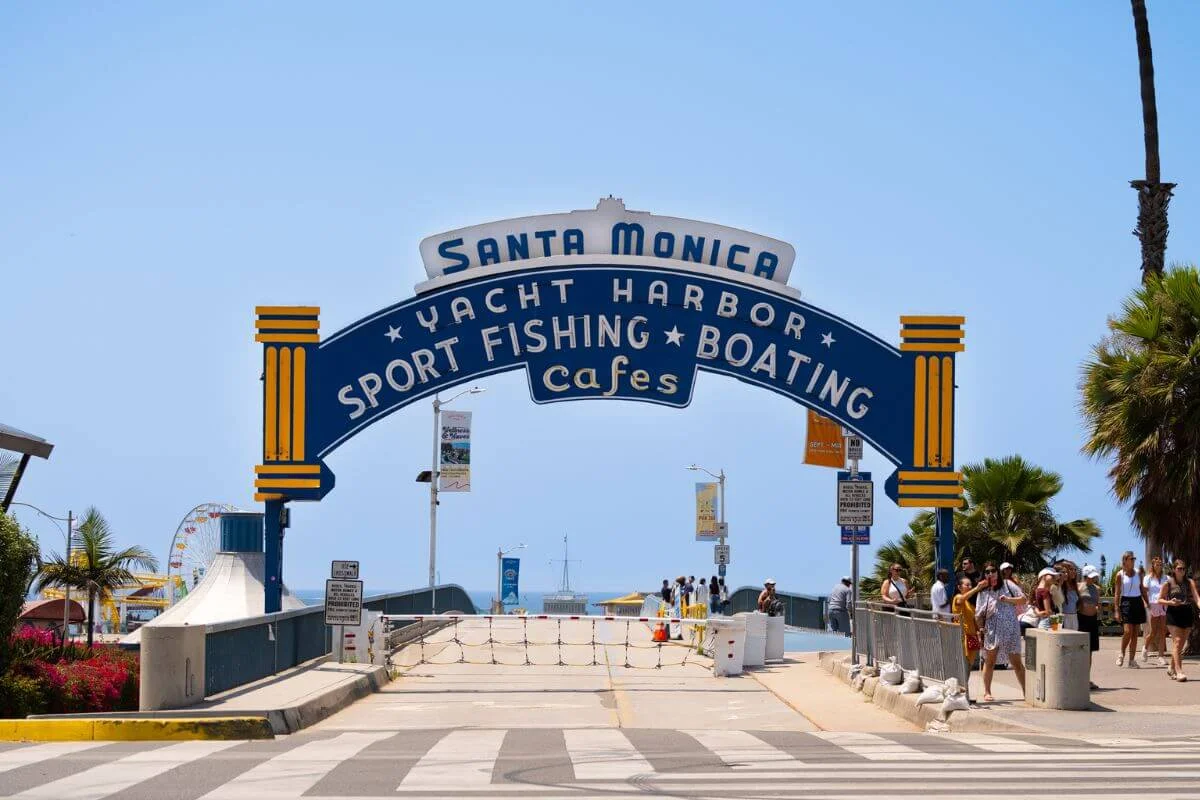 The iconic blue and yellow “Santa Monica Yacht Harbor Sport Fishing Boating Cafes” sign arches over the walkway leading to the Santa Monica Pier, with palm trees and crowds of visitors on a sunny day.