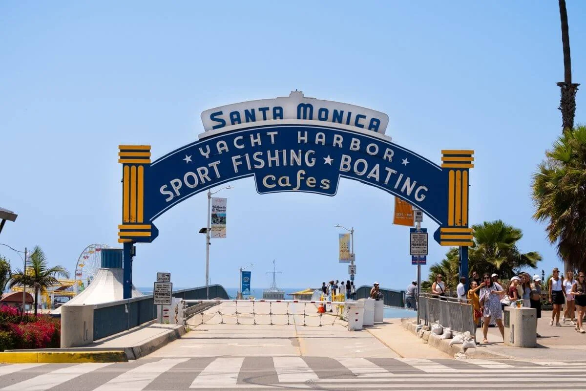 The iconic Santa Monica Pier entrance sign reading “Santa Monica Yacht Harbor Sport Fishing Boating Cafes,” with visitors walking nearby, palm trees on either side, and the ocean visible in the distance.