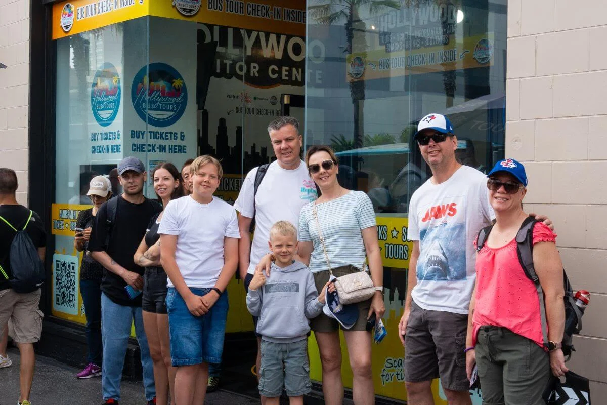 A group of smiling people, including adults and children, stand in front of the Hollywood Bus Tours visitor center entrance, ready to check in for their tour.
