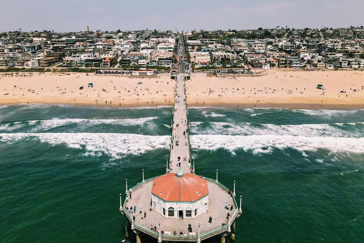 Drone view of a long pier stretching out into the ocean toward a round pavilion, with waves crashing below and a wide sandy beach and busy coastal neighborhood in the background.