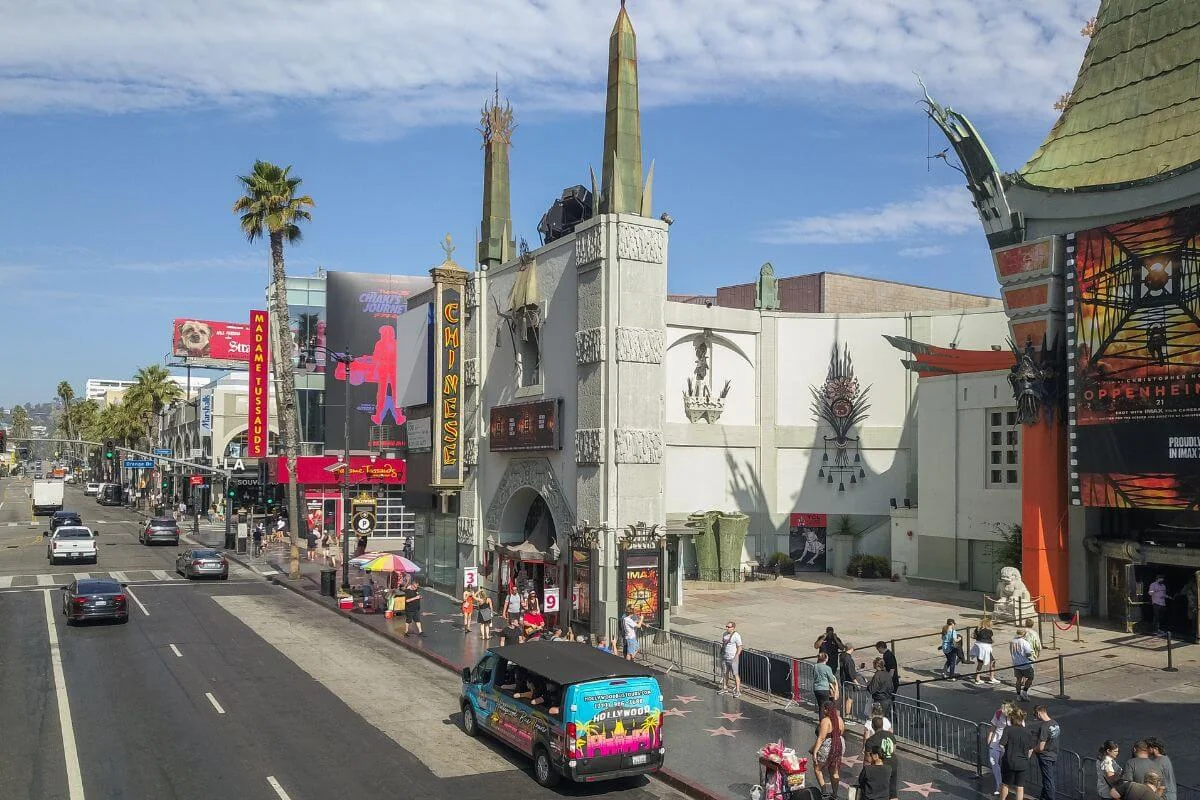A colorful Hollywood Bus Tours van drives along Hollywood Boulevard in front of the TCL Chinese Theatre, with people walking on the Walk of Fame and nearby attractions like Madame Tussauds visible on a sunny day.