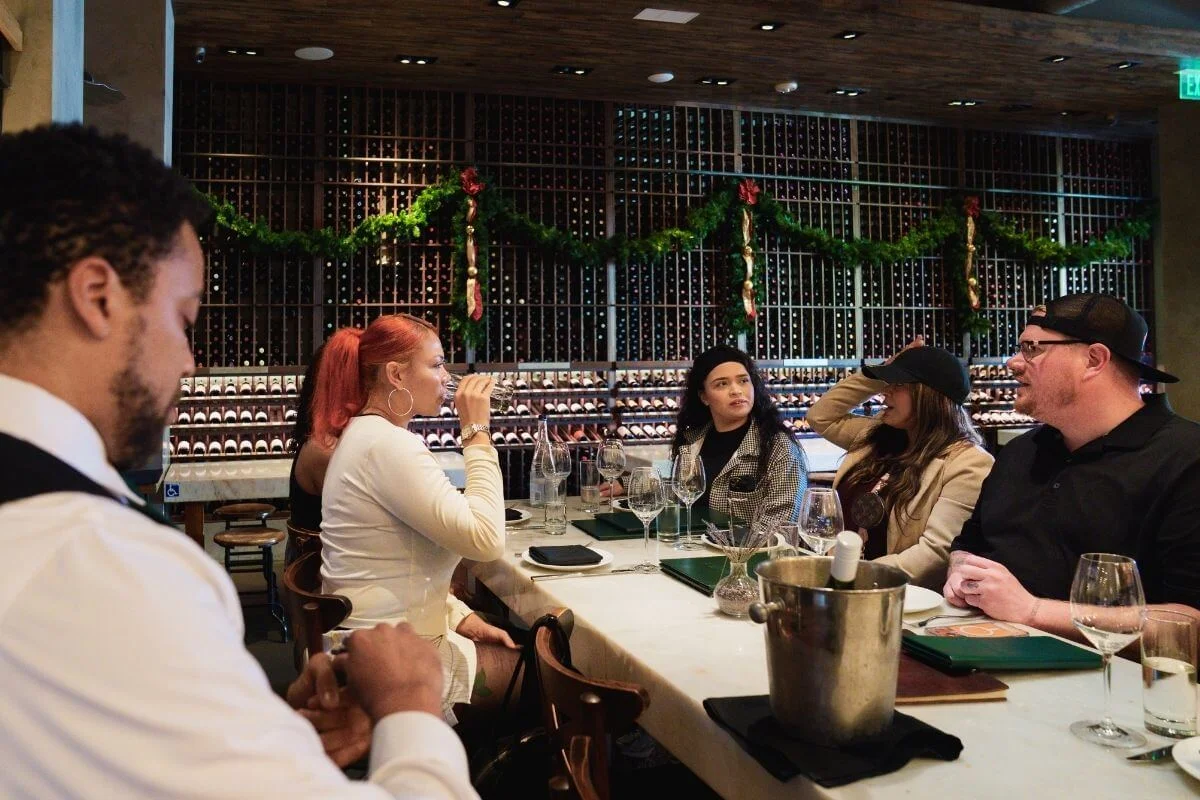 Group of people seated at a wine bar table with glasses and menus, talking while one person sips wine; wine racks and holiday garland in the background.