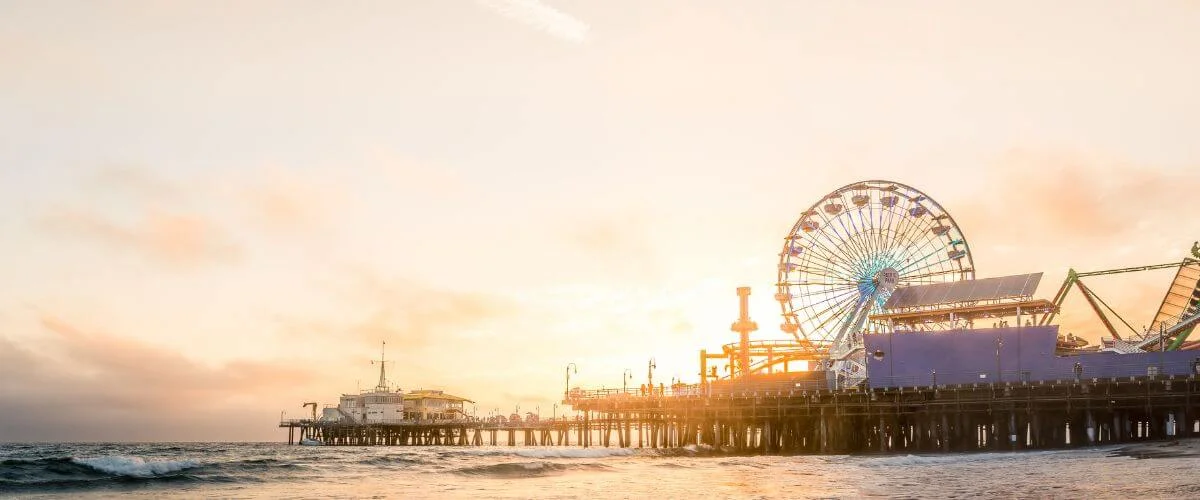 The Santa Monica Pier with a brightly lit Ferris wheel extending over the ocean at sunset, waves in the foreground and a warm golden sky in the background.