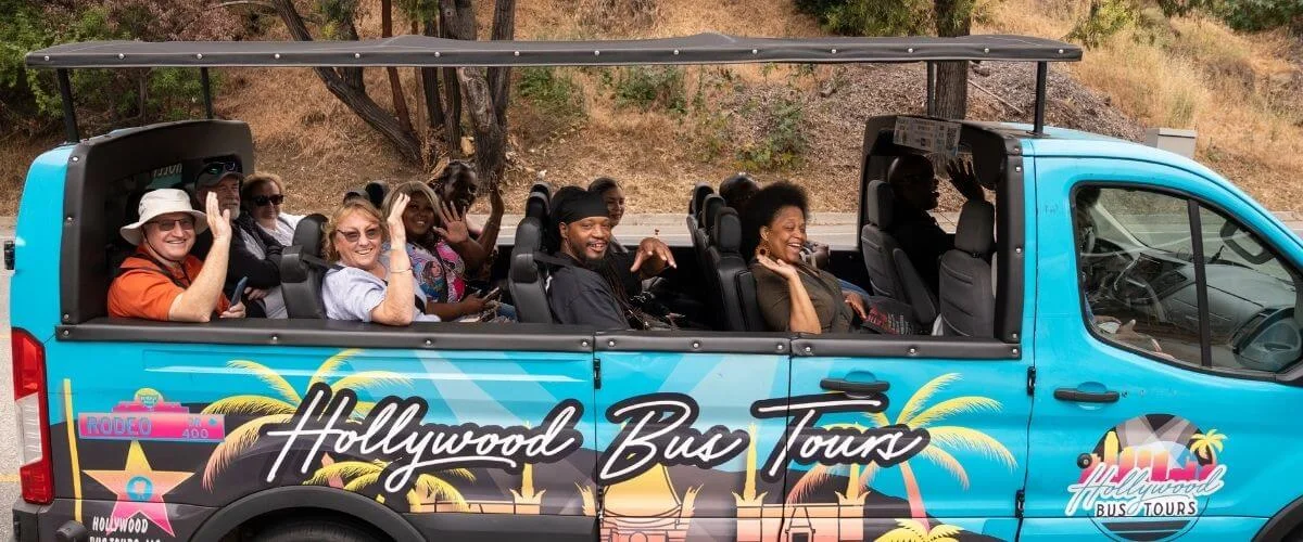 A bright blue open-air Hollywood Bus Tours van drives along the road, filled with smiling passengers waving at the camera against a backdrop of trees and hillside.
