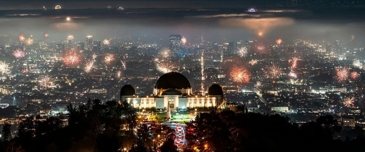 Night view of Griffith Observatory lit up on a hillside with the Los Angeles skyline in the background and fireworks bursting across the city under a hazy sky.