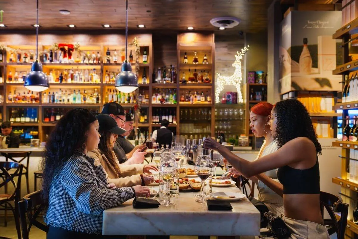 A group of friends sits at a marble table inside a warm, well-lit bar with shelves of bottles behind them, enjoying food and drinks during an evening meal.