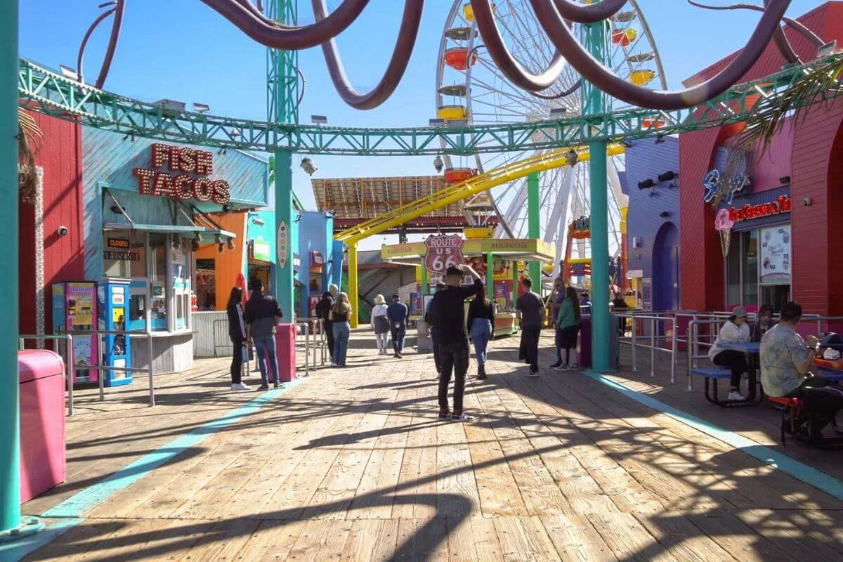 People walk along the wooden boardwalk at the Santa Monica Pier, passing bright food stands and shops, with a large Ferris wheel and roller coaster in the background on a sunny day.