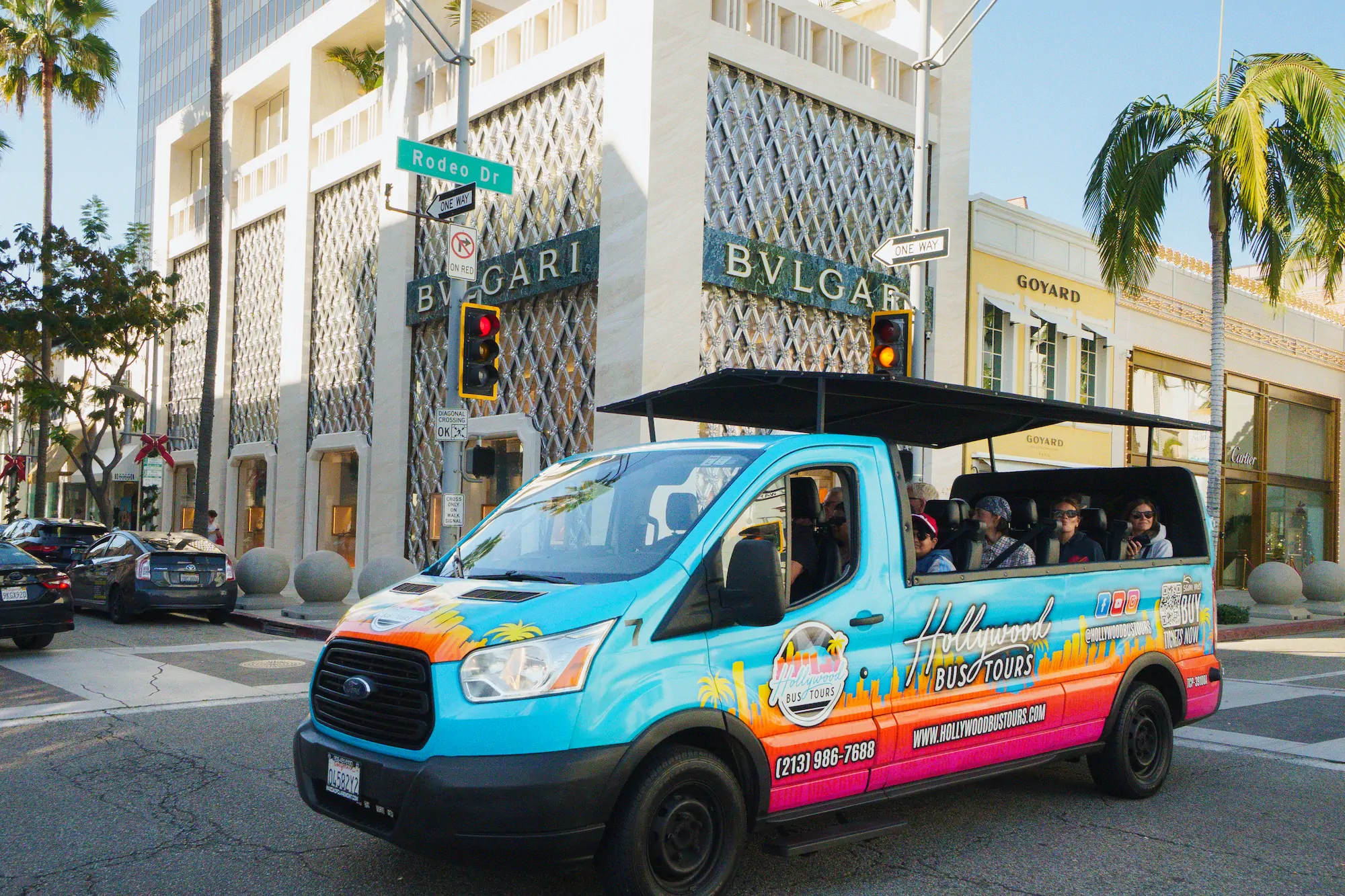A bright blue and orange open-air Hollywood Bus Tours vehicle filled with smiling passengers waves at the camera while driving along a tree-lined street.