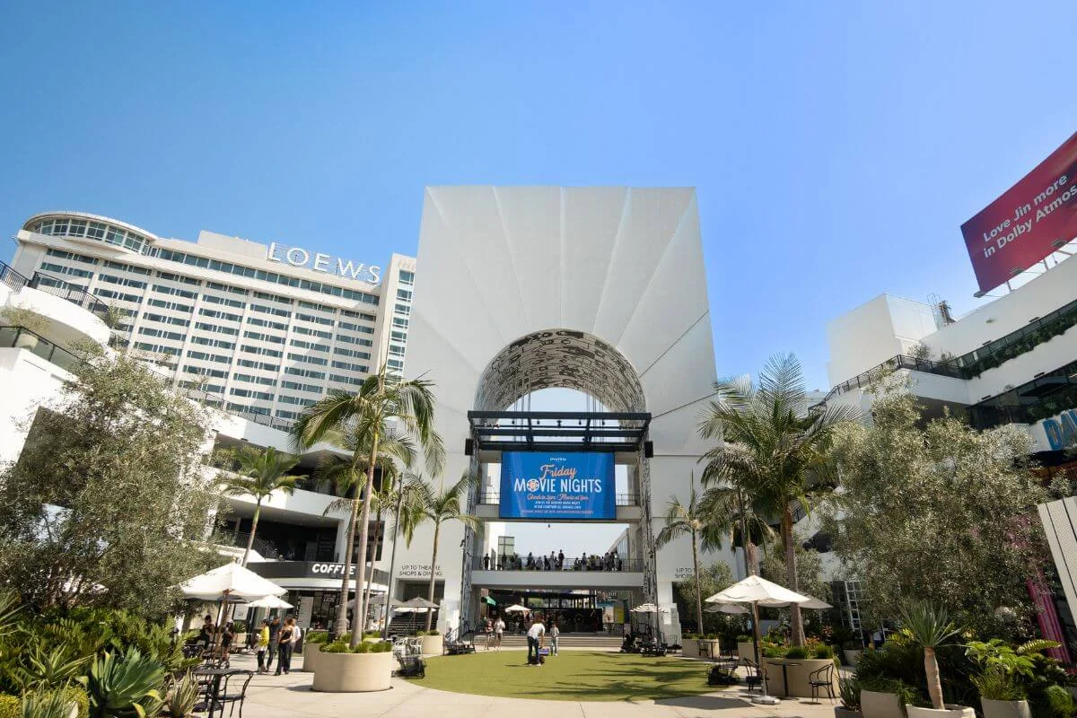 People walk and relax in a sunny outdoor courtyard with palm trees, café tables, and a large white archway displaying a “Friday Movie Nights” screen, with the Loews Hollywood Hotel rising in the background.