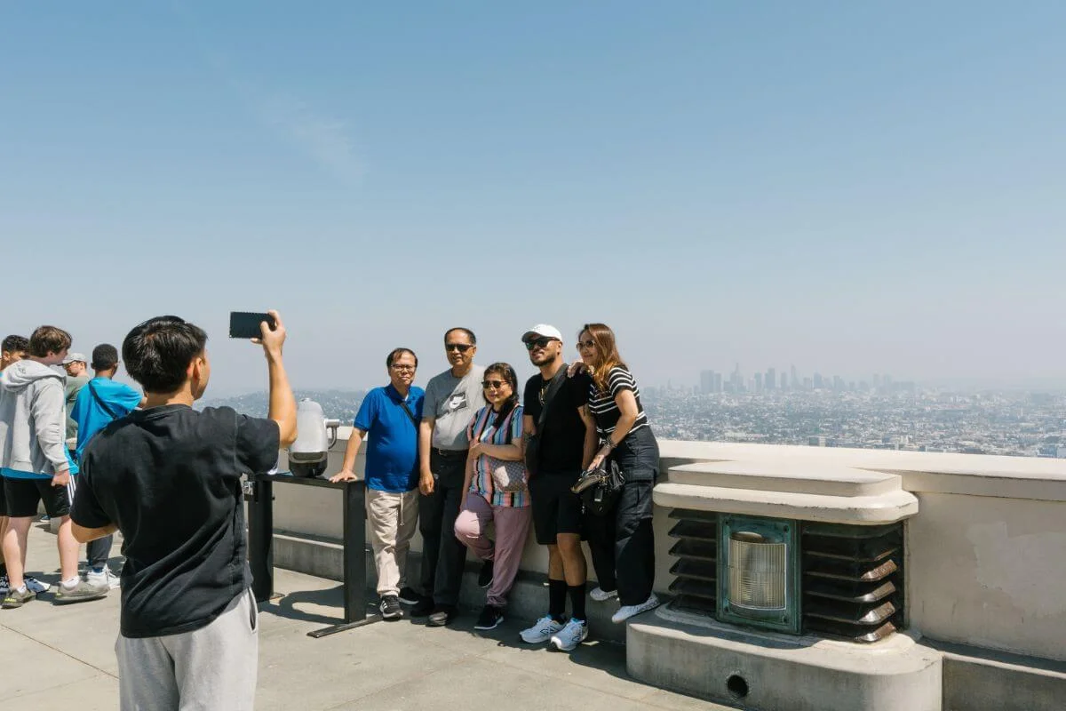 A person holds up a phone to photograph a group of five people posing along the Griffith Observatory terrace railing, with a hazy view of the Los Angeles skyline spread out in the distance under a clear blue sky.