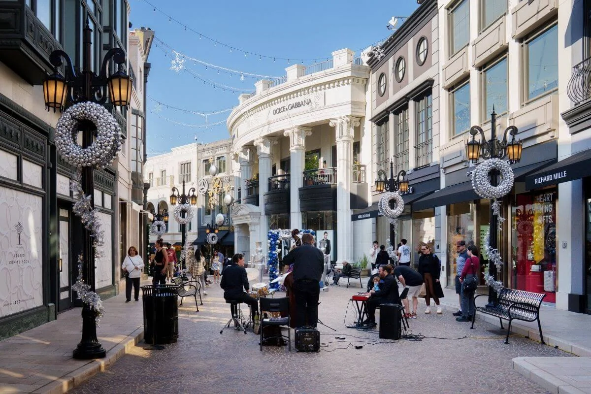 Pedestrian shopping street lined with luxury storefronts and holiday wreaths, with a small live band playing while people stroll and watch.