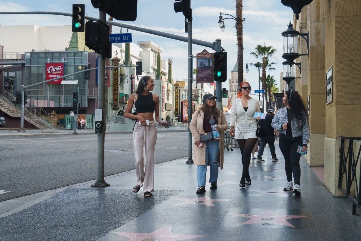 Four people walk along the Hollywood Walk of Fame on a sunny day, holding tour maps and chatting, with palm trees, traffic lights, and Hollywood Boulevard buildings in the background.