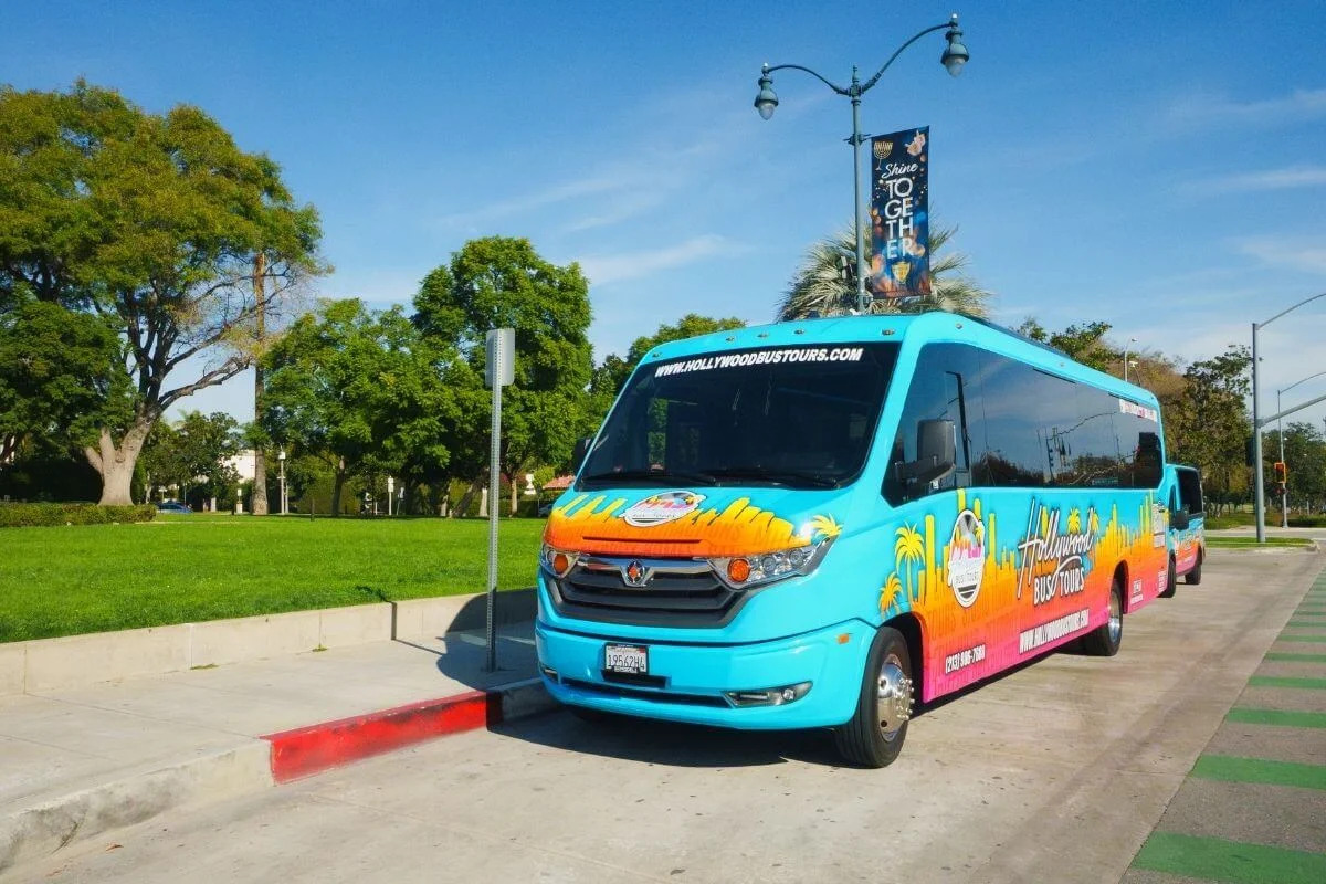 Bright turquoise Hollywood Bus Tours vehicle parked along a curb beside a grassy park with trees under a clear blue sky.