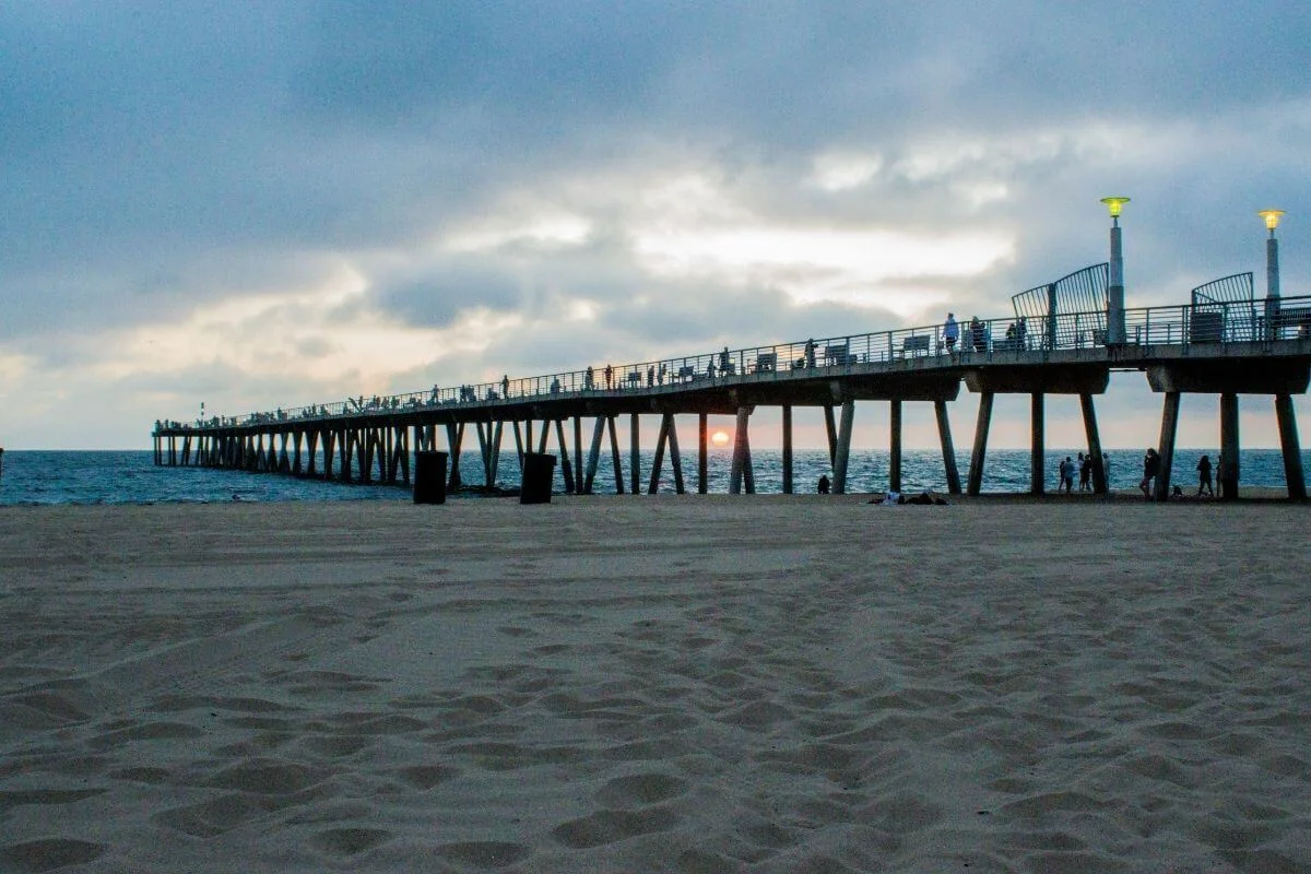 Long seaside pier extending over the ocean at sunset, with people walking along the pier, calm waves, sandy beach in the foreground, and cloudy evening sky.