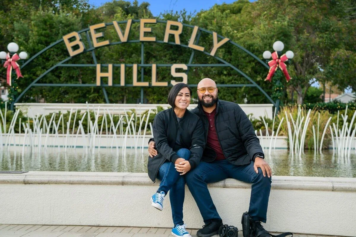 Smiling couple sitting together in front of the Beverly Hills sign and fountain, surrounded by greenery, with holiday decorations visible in the background.
