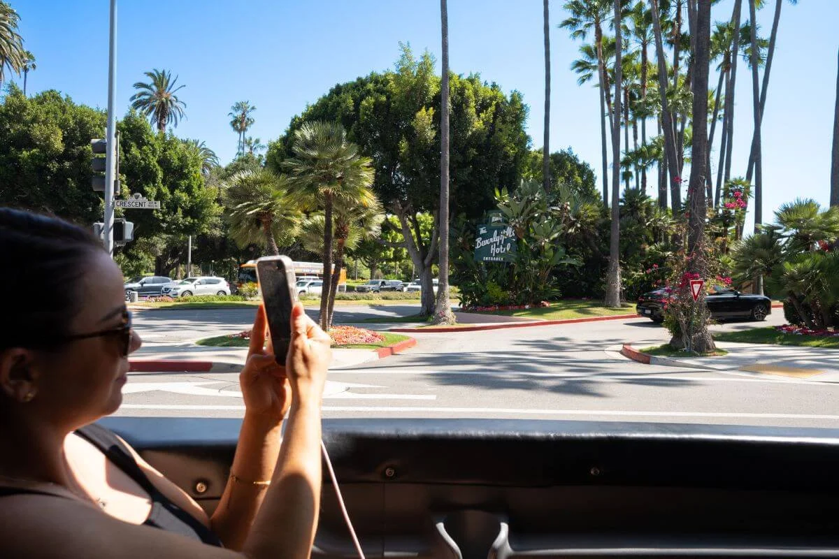 Person taking a photo from a vehicle of the Beverly Hills Hotel entrance, surrounded by palm trees and greenery on a sunny day.
