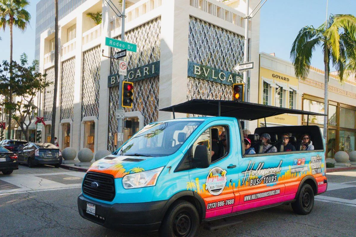 Colorful Hollywood bus tour driving past luxury stores on Rodeo Drive in Beverly Hills, with the BVLGARI storefront and palm-lined street visible.
