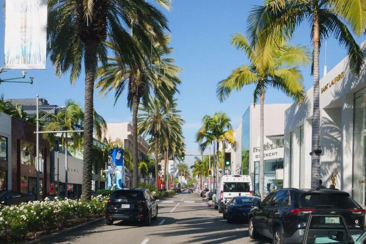 A sunny view of a palm-lined shopping street in Beverly Hills with luxury storefronts, cars driving along the road, and clear blue skies.