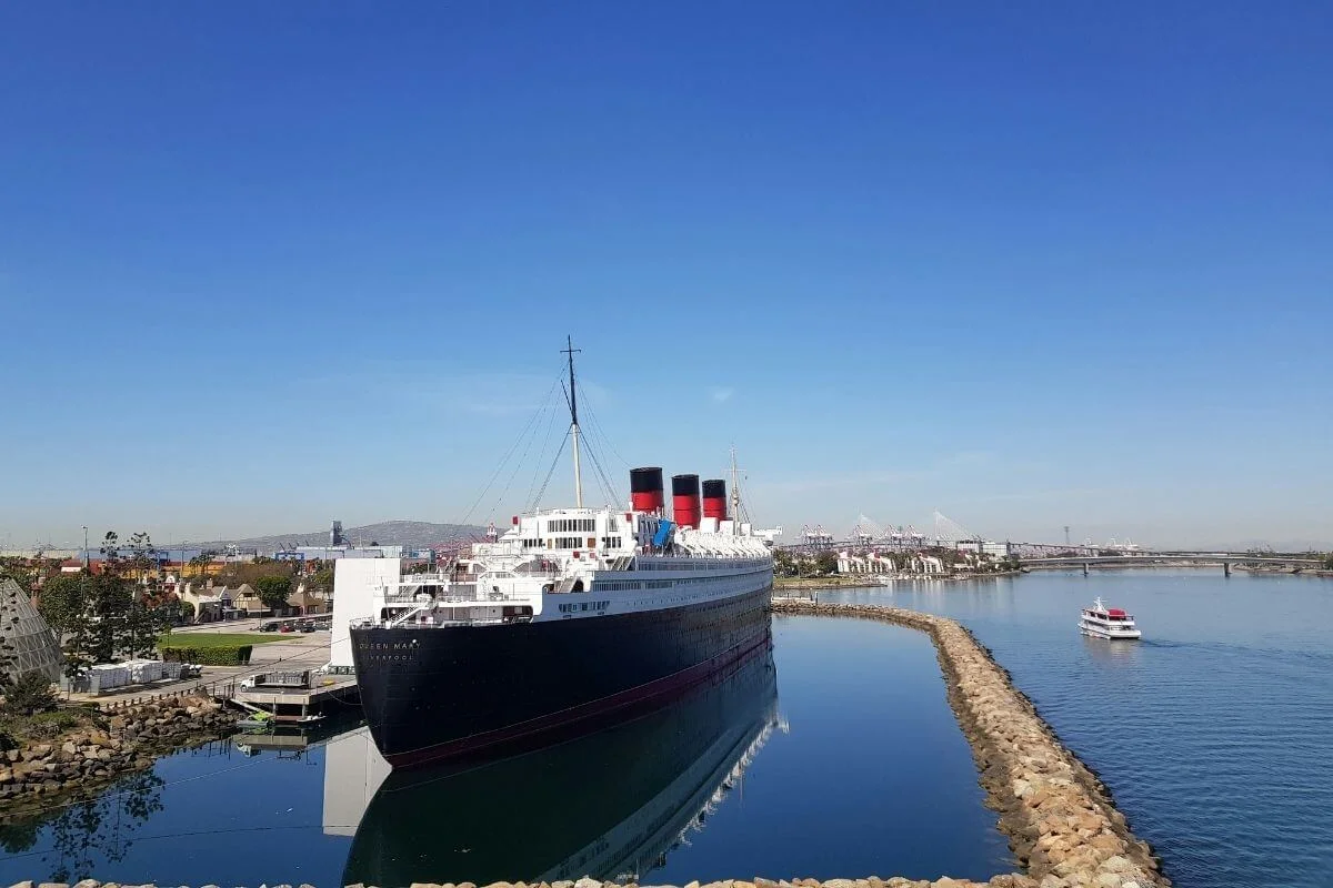 The Queen Mary ocean liner docked in Long Beach Harbor with calm water reflections and a small boat passing nearby under a clear blue sky