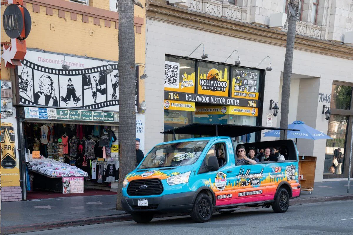 Colorful Hollywood Bus Tours van parked outside the Hollywood Visitor Center with passengers seated inside and souvenir shops nearby on Hollywood Boulevard