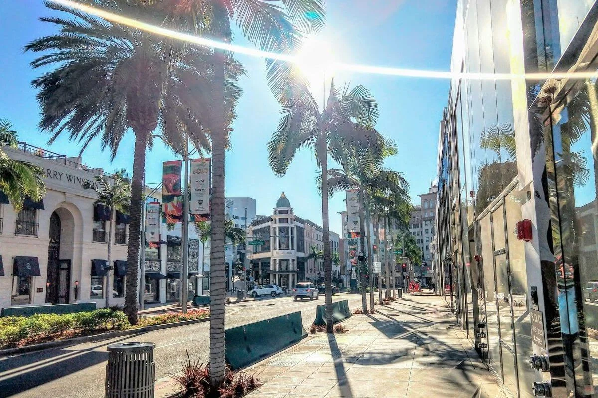 Sunny view of Rodeo Drive in Beverly Hills with palm trees, luxury storefronts, and light traffic along the street