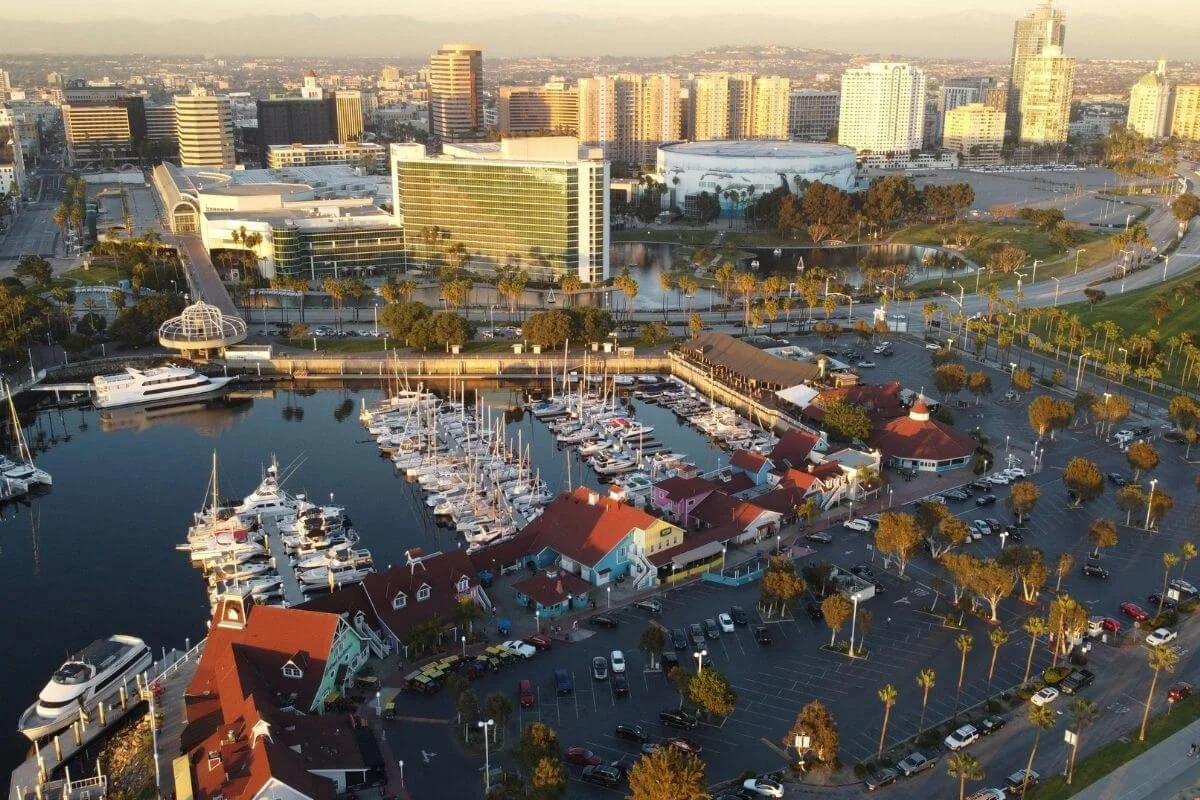Aerial view of Long Beach marina with docked boats, waterfront buildings with red roofs, and downtown skyline in the background during golden hour.