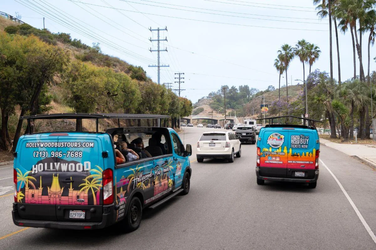 Two Hollywood Bus Tours vans carrying passengers drive along a palm-lined Los Angeles street during a city tour