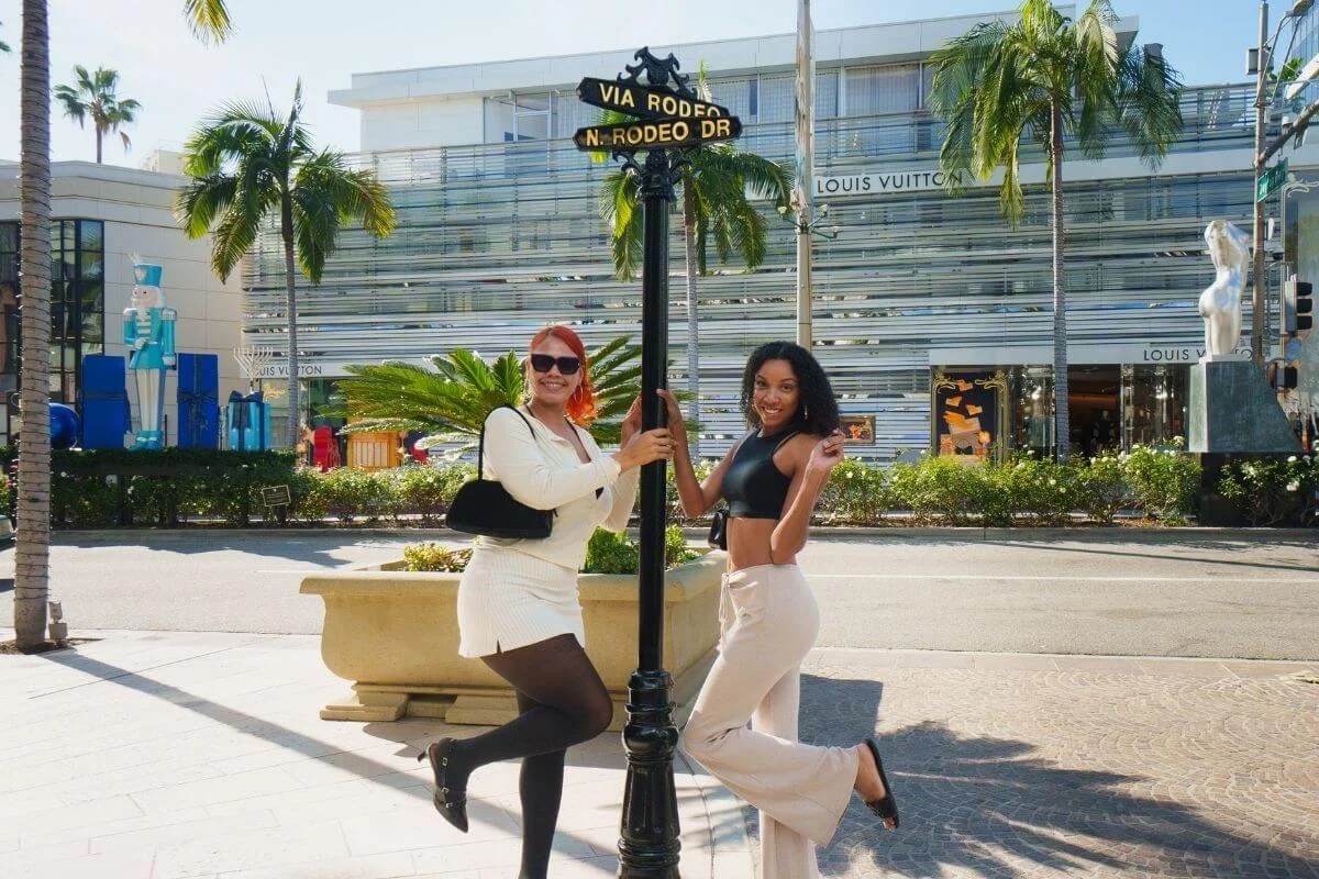 Two visitors posing by the Via Rodeo Drive and N Rodeo Drive street sign in Beverly Hills with luxury shops and palm trees in the background